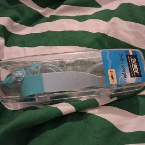 Swimpy blå simglasögon Junior - Snygga blå simglasögon från Swimpy för juniorer. De har en bred, justerbar rem i ljusblått och turkost, samt klara linser med transparenta detaljer. Perfekt för dig som vill ha stil på badhuset eller stranden.