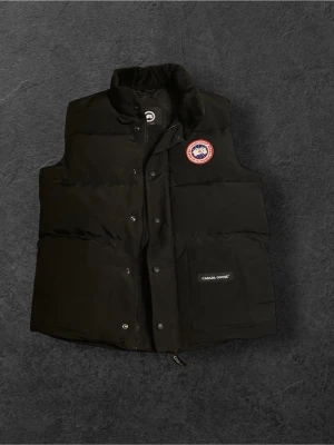 Canada Goose Väst - Svart Väst från Canada Goose med klassisk logotyp på bröstet. Västen har hög krage, tryckknappar och dragkedja framtill samt en praktisk ficka med märkets namn. Materialet är slitstark polyester och dunfyllning som håller dig varm under kyliga dagar.