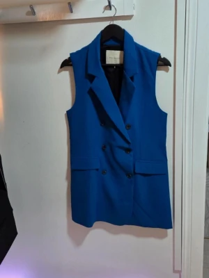 Free/Quent Elegant Väst / Ärmlös Kavaj Knallblå Stl S - En elegant och modern ärmlös kavaj/väst i en iögonfallande koboltblå färg. Plagget har dubbelknäppning (double breasted) och slag, vilket ger en sofistikerad look. Bär den över en skjorta, en tunn tröja eller direkt över en topp/linne på sommaren.  Märke: FREE/QUENT (Brands of Scandinavia)  Modell: FQKITA-WAISTCOAT Storlek: Small (S) Färg: Knallblå/Koboltblå                                   Skick: Aldrig använd. Säljes i nyskick.