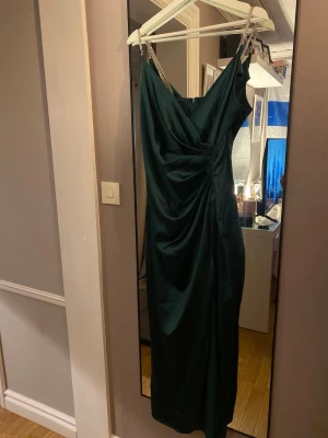 Mörkgrön långklänning med pärlband - Elegant mörkgrön långklänning med draperad framsida och smala axelband dekorerade med pärlor. Klänningen har en v-ringad urringning och figurnära passform som framhäver siluetten. Perfekt för speciella tillfällen.