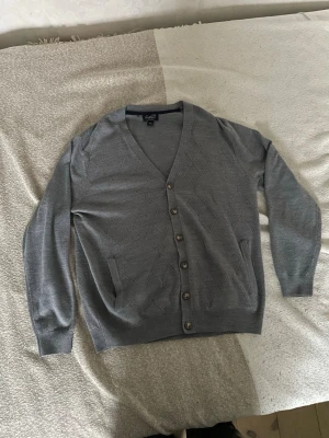 Grå cardigan med knappar - Stilren grå cardigan från Arnold Palmer med v-ringning och knäppning framtill. Tröjan har diskret rutmönster, två fickor och ribbade muddar vid ärmslut och nederkant. Perfekt för lager på lager och enkel att matcha.