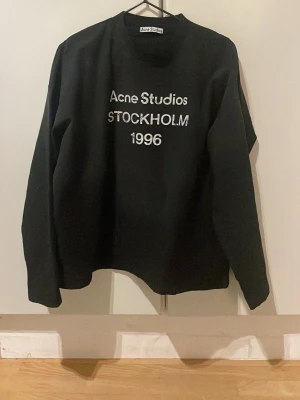 Acne studios tröja - Säljer en fet svart sweatshirt från Acne Studios med vit text 'Acne Studios STOCKHOLM 1996' på bröstet. Den är i mycket bra skick, storlek s. Personen på bilden är ca 180 och väger ca 70 kg. Skriv vid minst funderingar!