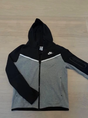 Nike grå och svart hoodie med dragkedja - Snygg hoodie från Nike i grått och svart med vit logga på bröstet. Hoodien har dragkedja framtill, huva och två fickor. Ärmarna är svarta och resten av tröjan är grå med en vit linje över bröstet. Perfekt för en sportig och avslappnad stil.