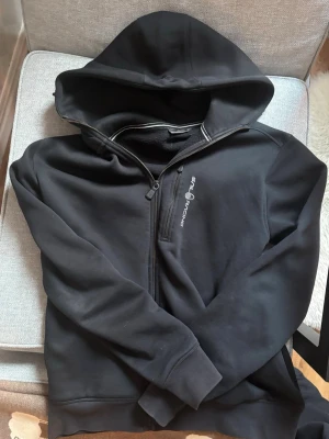 Svart hoodie från Sail Racing - Svart hoodie från Sail Racing med dragkedja och broderad logga på bröstet. Mjuk insida och klassisk huva. Perfekt för dig som gillar stilren och sportig design. Tillverkad i ett tjockare material som känns riktigt skönt mot huden.