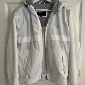 Vit vindjacka från Hackett London S - Snygg vit vindjacka från Hackett London Heritage med grå huva och dragkedja framtill. Jackan har två fickor med lock, ribbade muddar och diskret logga på bröstet. Perfekt för dig som gillar stilren och sportig look.