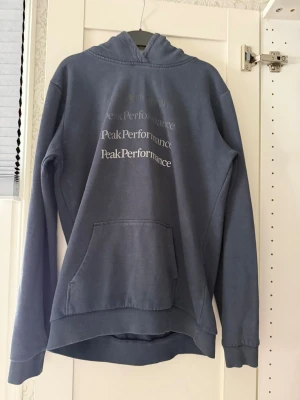 Blå hoodie från Peak Performance - Mysig blå hoodie från Peak Performance med tryckt logga i vitt på bröstet. Hoodien har en stor känguruficka framtill, ribbade muddar vid ärmslut och nederkant samt en klassisk huva. Perfekt för en avslappnad och sportig stil.