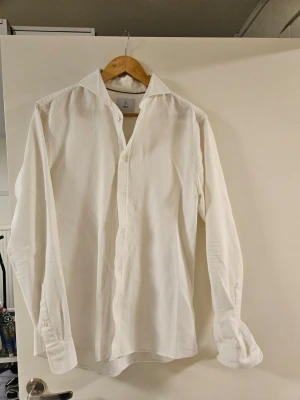 Vit långärmad skjorta  - Klassisk vit skjorta från Bläck med lång ärm och knappar framtill. Skjortan har en stilren krage och är tillverkad i ett lätt, strukturerat bomullstyg som ger en fräsch och clean look. Perfekt för dig som gillar enkel och tidlös stil.