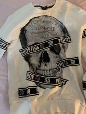 Vit sweatshirt med dödskalle Philipp Plein - Cool vit sweatshirt från Philipp Plein med stort dödskallemotiv i svart och silver på framsidan. Tröjan har svarta band med texten 'PHILIPP PLEIN 1978' och ribbade muddar. Perfekt för dig som gillar streetwear och statementplagg.