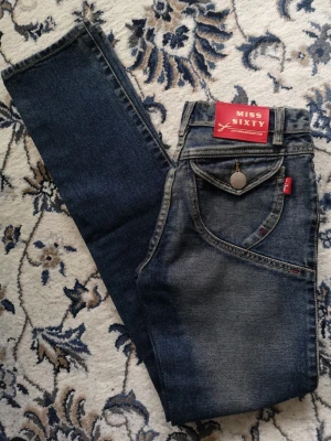Mörkblå jeans från Miss Sixty - Snygga mörkblå jeans från Miss Sixty med klassisk femficksmodell och unika sömdetaljer vid fickorna. Jeansen har normal passform och raka ben. Materialet är slitstarkt denim i bomull. Perfekta för dig som gillar en tidlös och cool look. stl 26 midje 68cm innerbenslängd 82 cm. längd 107cm. Använts ett fåtal gånger, i finfin skick, djur och rökfritt hem.