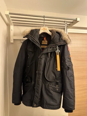 Svart Parajumpers parkas med pälskrage - Säljer en svart Parajumpers parkas med avtagbar pälskrage, flera fickor och ikonisk gul rem vid halsen. Jackan har dragkedja och knappar framtill samt lång ärm. Perfekt för kalla dagar och riktigt snygg streetstil. Äkta såklart, och använd enbart 1 vinter.