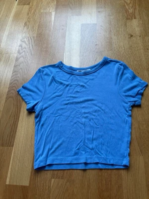 Blå croppad t-shirt från Zara - En enkel och stilren blå croppad t-shirt från Zara . T-shirten har rund halsringning och korta ärmar. Perfekt till jeans eller kjol för en avslappnad look. Säljer på grund av för liten storlek!