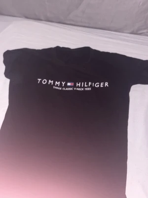 Svart v-ringad t-shirt Tommy Hilfiger - Svart t-shirt från Tommy Hilfiger med v-ringning och tryckt logga på bröstet. Klassisk och enkel design som passar till det mesta. Tillverkad i mjuk bomull för skön känsla hela dagen.