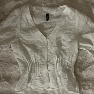 Vit blus med v-ringning från H&M - Säljer en fräsch vit blus från H&M med v-ringad hals och knappar framtill. Blusen har lång ärm och snygga rynkade detaljer vid midjan som ger en feminin siluett. Perfekt för dig som gillar stilrena och enkla plagg!