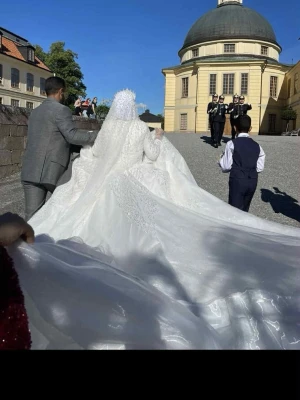 Vit lång bröllopsklänning med slöja - Elegant vit bröllopsklänning med lång, vid kjol och vackra spetsdetaljer. Klänningen har långärmade ärmar, hög hals och matchande slöja. Materialet är glittrigt och skimrande, vilket ger en lyxig känsla. Perfekt för dig som vill ha en klassisk och pampig look.