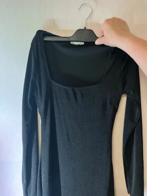 Svart långärmad klänning Nly trend - En stilren svart klänning med lång ärm och fyrkantig ringning. Klänningen har en kroppsnära passform och är tillverkad i ett mjukt, stretchigt material som ger en snygg siluett. Perfekt för dig som gillar minimalistisk och t