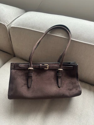 Mörkbrun Väska H&M - En avlång, coatad axelremsväska med regelbar axelrem, aldrig använd.  Höjd 17 cm  Bredd 31 cm Djup 11 cm