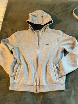 Lacoste zip tröja - En fin Lacoste sport zip i grå färg med en mörkblå hoodie. Tröjan är i storlek 160 men är lite mindre i storlek( kanske passar 150-160). Det är inga defekter på tröjan men lite text under taggen se bild 4. Skriv för fler frågor!