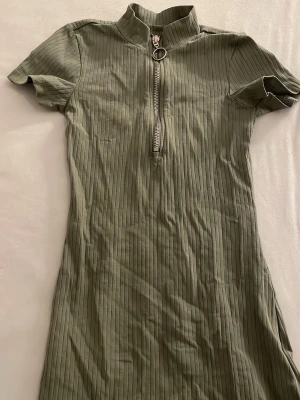 Grön ribbad kortklänning Sinsay - Snygg grön ribbad kortklänning från Sinsay med halv dragkedja och ringdetalj framtill. Klänningen har korta ärmar och en hög krage som ger en cool look. Perfekt för dig som gillar en enkel men trendig stil.