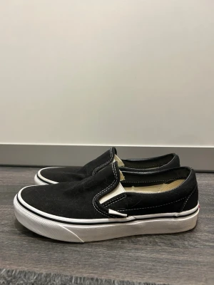 Svarta slip-on sneakers från Vans - Klassiska svarta slip-on sneakers från Vans med vit sula och kontrasterande vita sömmar. Skorna har en låg profil och är tillverkade i canvas med elastiska paneler på sidorna för enkel på- och avtagning. Perfekta för en avslappnad och stilren look.