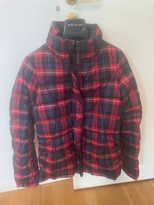 Rutig pufferjacka från River Island - Snygg mysig piffig lätt vikts jacka från Tomy Hilfinger i rött och mörkblått i klassiskt rutigt mönster. Jackan har hög krage, quiltad design och dragkedja framtill. Perfekt för kalla dagar. Tillhörande påse om man vill ta med sig jackan och ta av den så tar den liten plats Jackan nästan aldrig använd. Säljes pga fel köp. 