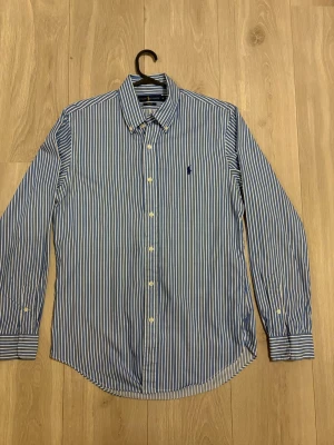 Blårandig skjorta från Ralph Lauren - Klassisk blårandig skjorta från Ralph Lauren med vit knappstängning och broderad logga på bröstet. Skjortan har lång ärm, krage och är tillverkad i bomull för en bekväm känsla. Perfekt för en stilren och tidlös look.