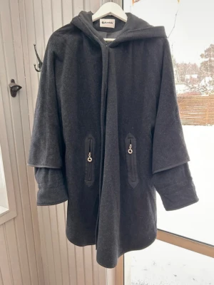 Mörkgrå kappa med huva i capemodell - Elegant yllekappa i cape-modell med huva och dekorativa detaljer. Den har en unik design med korta ärmar och praktiska fickor med dragkedja. Kappan knäpps med dolda knappar. Kappan är gjord i ull och kaschmir.  Mycket bra skick.