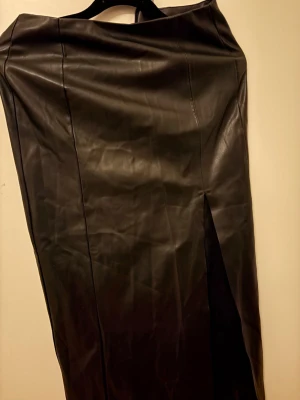 Svart lång skinnkjol med slits - Snygg svart långkjol i skinnimitation med markerade sömmar och en stilren slits framtill. Kjolen har en rak passform och ger en cool och trendig look. Perfekt för dig som gillar att sticka ut med en edgy stil.