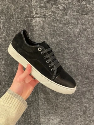 Svarta sneakers från Lanvin - Stilrena svarta sneakers från Lanvin med ovandel i mocka och detaljer i blankt skinn. Skorna har vita sulor, svarta skosnören och metallfärgade öljetter. Perfekta för dig som vill ha en clean och lyxig look.