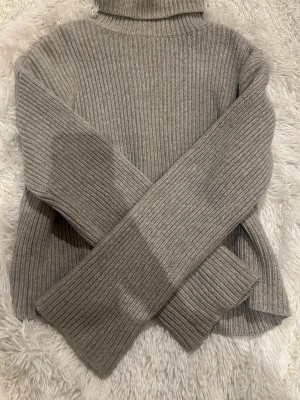 Grå ribbstickad polotröja - Säljer en stilren grå ribbstickad polotröja med hög krage och långa ärmar. Tröjan har en klassisk rak passform och är gjord i ett mjukt, tjockare material som håller dig varm. Perfekt för kyliga dagar och enkel att matcha med olika outfits.