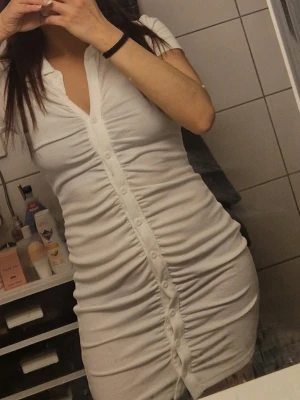 Vit kortärmad knappklänning - Säljer en vit kort klänning med knappar framtill och v-ringning. Klänningen har korta ärmar och är figurnära med snygga rynkade detaljer längs knappraden. Perfekt för dig som gillar enkel men stilren design.