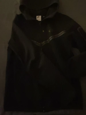 Svart Nike Tech - Säljer en svart hoodie från Nike med dragkedja framtill och klassisk huva. Hoodien har en diskret Nike-logga på bröstet och en snygg detalj med ett band tvärs över bröstet. Tillverkad i mjukt bomullsmaterial, perfekt för en avslappnad streetwear-look. Säljer eftersom den har blivit för liten för mig