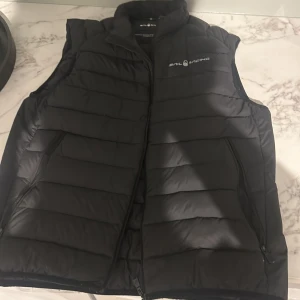 Svart dunväst från Sail Racing - Svart dunväst från Sail Racing med quiltad design och hög krage. Västen har dragkedja framtill och två sidofickor med dragkedja. Märkets logga syns på bröstet och nacken. Materialet är polyester och dun, perfekt för kyliga dagar.