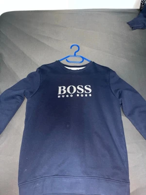 Mörkblå sweatshirt från Hugo Boss - Snygg mörkblå sweatshirt från Hugo Boss med stor vit logga på bröstet. Klassisk rund halsringning och mjukt material som känns skönt mot huden. Perfekt för en clean och stilren look.
