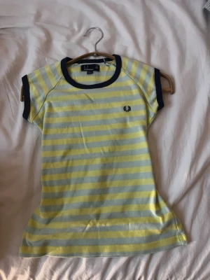 Y2K vintage Randig t-shirt från Fred Perry - Gul och ljusblå randig t-shirt från Fred Perry med mörkblå detaljer vid ärmslut och halsringning. Klassisk logga broderad på bröstet. Skön passform och perfekt till sommaren. Tillverkad i mjuk bomull.