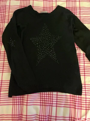 Svart tröja med stjärndetaljer från Vila - Svart långärmad tröja från Vila med en stor stjärna av små nitar på bröstet och en mindre stjärna på ena ärmen. Tröjan har rund halsringning och är i mjukt material, perfekt för en chill och snygg look.