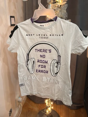 Vit t-shirt med hörlursprint - Vit t-shirt från max med tryck av stora hörlurar och texten 'THERE'S NO ROOM FOR ERROR' i lila. T-shirten har rund hals med lila kant och svarta detaljer på axlarna. Perfekt för dig som gillar gaming och streetstyle.