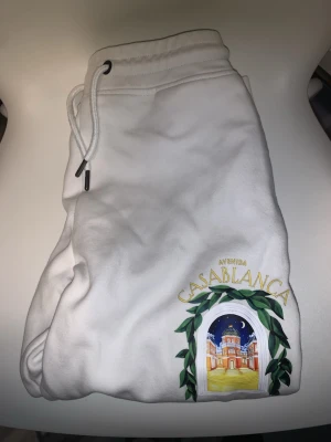 Vita mjukisbyxor från Casablanca - Säljer ett par vita mjukisbyxor från Casablanca med snyggt tryck vid benet som föreställer en byggnad och gröna blad. Byxorna har dragsko i midjan och är perfekta för en chill och avslappnad stil.