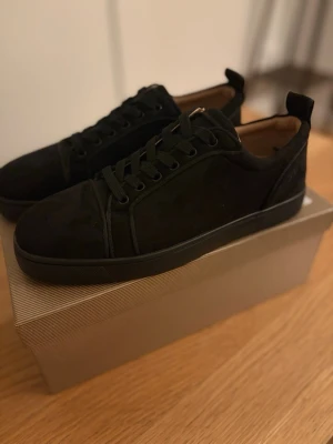 Svarta sneakers från Christian Louboutin - Stilrena svarta sneakers från Christian Louboutin med klassisk röd sula. Skorna har snörning och är tillverkade i mocka med diskreta detaljer. Perfekta för dig som vill ha en clean look med exklusiv känsla.