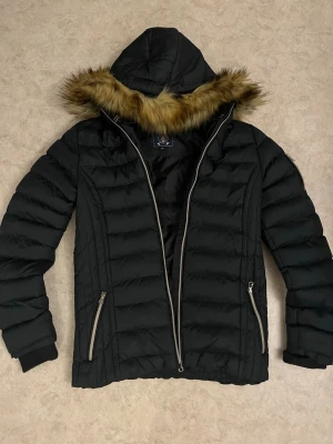 Svart dunjacka från Vinson Polo Club - Svart dunjacka från Vinson Polo Club med huva och fejkpäls runt kragen. Jackan har quiltad design, två dragkedjefickor och en snygg patch på ärmen. Perfekt för kalla dagar och riktigt stilren look.