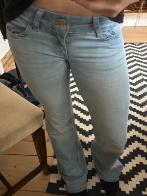 Ljusblå bootcut jeans med broderi - Säljer ett par ljusblå bootcut jeans med snygga röda broderade detaljer på bakfickorna och dubbla knappar i midjan. Jeansen har en klassisk femficksmodell och är tillverkade i mjukt denimtyg. Perfekta för dig som gillar en avslappnad men trendig look.