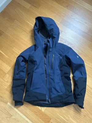 Mörkblå skidjacka med huva Peak Performance  - Snygg mörkblå skidjacka från Peak Performance med huva och dragkedja framtill. Jackan har en bröstficka med dragkedja och svarta paneler längs sidorna och ärmarna. Perfekt för blåsiga dagar och outdoor-aktiviteter. Snölås inuti. Jackan är i mint condition använd 2 gånger. 