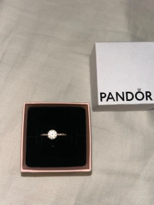 Guldfärgad ring med sten från Pandora - Snygg guldplätterad ring från Pandora med en stor, gnistrande klar sten i mitten och mindre stenar längs bandet. Ringen är knappt använd och nypris är 1199kr. Kommer i original ask.