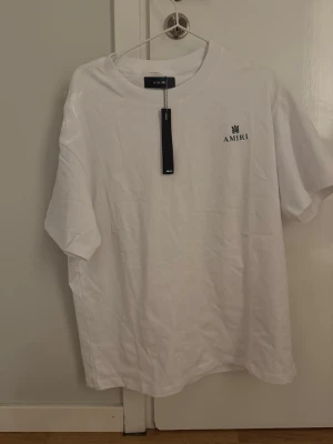 Vit t-shirt från Amiri med logga - Vit t-shirt från Amiri med svart logga på bröstet och en diskret detalj på ryggen. Klassisk rund hals och korta ärmar. Tillverkad i mjuk bomull för en skön känsla och stilren look. Storlek M. Helt oanvänd. Kan gå ner i pris på snabb affär