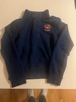 Blå Hollister hoodie med knappar - Mörkblå hoodie från Hollister med hög krage och tre knappar framtill. Broderad logga med texten 'Hollister So Cal' och en röd mås på bröstet. Stor ficka framtill och ribbade muddar vid ärmslut och midja. Perfekt för en avslappnad och cool stil.