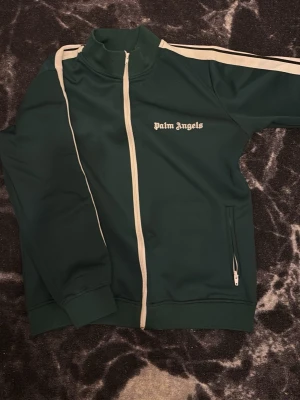 Grön track jacket från Palm Angels - Mörkgrön track jacket från Palm Angels med vita ränder längs ärmarna och vit dragkedja. Jackan har hög krage, två fickor med dragkedja och Palm Angels-logga på bröstet. Tillverkad i polyester för en sportig och bekväm känsla.
