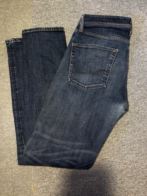 Jack & Jones blå jeans comfort fit - Snygga blå jeans från Jack & Jones med comfort fit och klassisk femficksdesign. Jeansen har knäppning med knappar, kontrastsömmar och en mörk tvätt med slitningar. Perfekta för dig som gillar en avslappnad men stilren look.