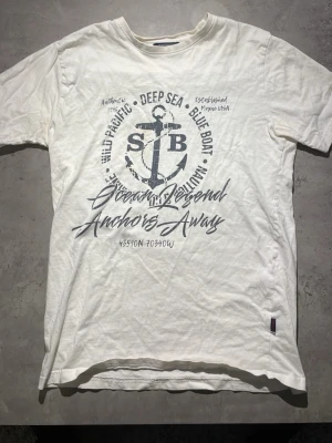 Vit Sebago t-shirt med ankare - Säljer en vit t-shirt från Sebago med ett stort tryck av ett ankare och marin text på framsidan. T-shirten har korta ärmar och är gjord i mjuk bomull. Perfekt för dig som gillar en avslappnad och nautisk stil.