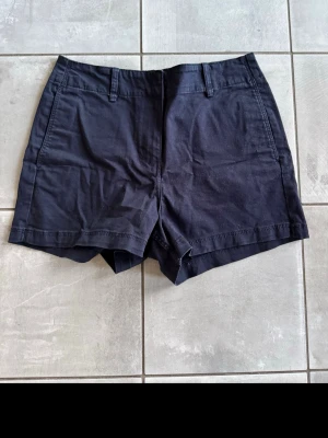 Blå shorts med fickor - Snygga svarta shorts med normal passform och praktiska fickor både fram och bak. Shortsen har hällor för bälte och är tillverkade i ett mjukt bomullstyg, perfekta för varma dagar. Klassisk design som funkar till det mesta.