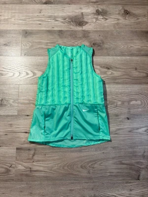 Nike Vest - Snygg turkos sportväst från Nike med quiltad framsida och två stora fickor framtill. Västen har hel dragkedja och är tillverkad i lätt, funktionsmaterial som passar perfekt för träning eller lager-på-lager. Diskret Nike-logga på ena fickan. Storlek M passar S