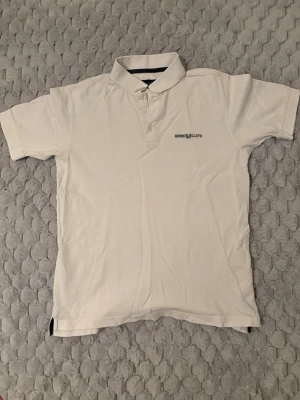 Vit pikétröja från Henri Lloyd - Klassisk vit pikétröja från Henri Lloyd med korta ärmar och krage. Tröjan har knappar framtill och en liten logga på bröstet. Perfekt till en clean och enkel stil.